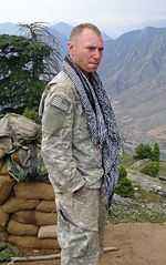 A man in his camoflouge military uniform wearing a scarf around his neck. He is looking at the camera and has his hands in his pockets. Several mountains are visible in the background.