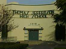 A photograph of a beige building with green lettering on the front reading "THOMAS JEFFERSON HIGH SCHOOL" above a set of green doors that are at the top of some stairs