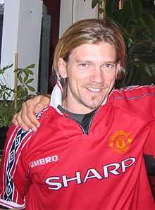 A photograph of a man with medium-length dirty blonde hair wearing a red shirt.