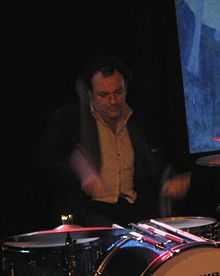 A 45-year-old man is shown at his drum kit. Both drum sticks are blurred in motion. Part of the drum kit is out of view. The man is dressed in a dark jacket with a light shirt, his eyes are focussed down. Behind him is a mostly dark background, past his left shoulder is a blue and white picture.