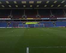 A two-tiered football stand with claret seats in the upper tier and light blue ones in the lower. In the lower tier, a group of people in fluorescent yellow jackets are congregated. Two sets of goalposts can be seen in front of the stand.