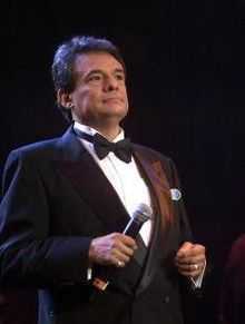 A man appears on a stage. He is wearing a black tuxedo.