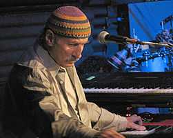 A man behind a microphone, with one of his hands on a black and white keyboard, wearing a dress shirt and a multi-colored cap.