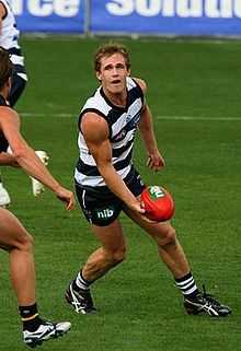 Young male athlete stands in a navy blue and white striped sleeveless shirt and blue navy shorts. With an opponent approaching him, he looks to the left and prepares to handpass the red football.