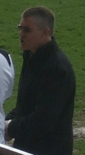 A man with grey hair is wearing a black jacket. He is standing on a grass field.