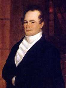 A man with dark, curly, receding hair wearing a high-collared white shirt and black jacket