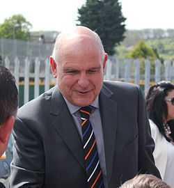 A balding man in a grey suit and a striped tie smiles as he greets Luton supporters at Kenilworth Road