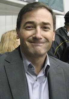 Upper body of a man.  He is wearing a grey suit coat over a light blue shirt.  He has short, dark hair and a wide grin.