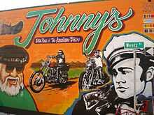 Wall of a building with mural of a bearded man with sunglasses wearing a beret, a young man in a white hat in front of two riders on chopper motorcycles below the legend Johnny's the birthplace of the American biker.