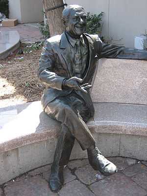 A statue seated on a curved bench, behind which is the trunk of a tree adjacent to the wall of a building.