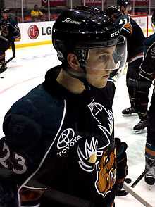 Young male ice hockey player, visible from waist up, skating while facing the right. He is wearing a helmet with a visor, and the uniform logo, a snarling moose, is partially visible on his jersey. In the background three of his teammates, partially obscured, are also warming up.