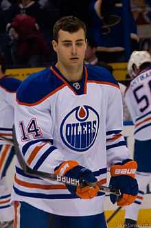 A Caucasian ice hockey player wearing a white jersey with a blue and orange circular logo with the word "OILERS".