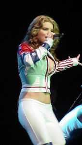 A woman is performing a song over the stage. She wears a white jacket and pants.