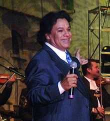 A man standing with black hair, smiling, wearing a blue jacket, white shirt and blue tie, holding a microphone in his right hand.