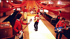 A seven member band are playing instruments against the backdrop of orange, yellow, and red drapes.