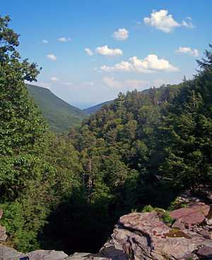 A view over a long, narrow valley with steep wooded sides. A rocky ledge is in the foreground