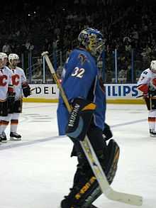 An ice hockey goaltender skates towards the right of the picture. He wears a blue jersey and blue helmet, with black leg pads and blocker