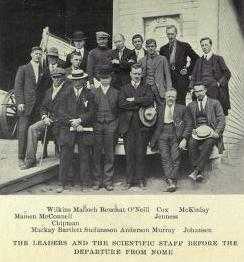 A group of 16 men, standing or sitting, on the deck of a ship with a small lifeboat visible, left background. The group's pose is casual and the men are variously attired, many in suits with casual hats of different sorts.