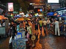 Night scene on a pedestrian street, with many people and street vendors; shops along the street bearing brightly lit signs with names like "99Fashion", "Brick Bar", "Mulligans Irish Bar", and Pepsi and McDonald's logos