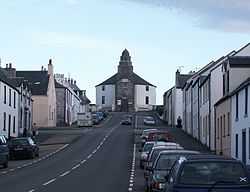 A wide street leads up a slope with parked cares and stone houses painted in whites and yellows on either side. At the end of the street there is a grey and white building with a short spire.