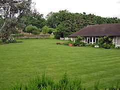 A white building with a black roof beside a large, well-kept lawn. Many trees are in the background of the picture, behind the building.