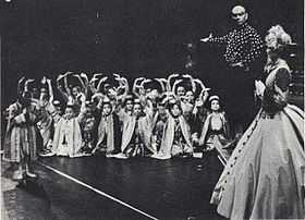 Black and white photo of a theatrical scene: a middle-aged man with a shaved head and imposing presence has his right arm extended to introduce a group of children in Asian dress to a woman in a crinoline dress and bonnet in the foreground at right, who is partially turned upstage.  The children are mostly kneeling and have their arms raised in greeting; one child (probably Crown Prince Chulalongkorn) stands and bows.