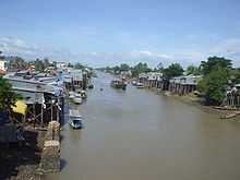 Brown-coloured waterway, with many small boats and elevated wooden and thatched houses visible. Foliage is also in abundance.