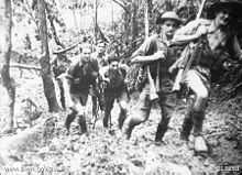 Soldiers in short sleeve shirts and shorts, slouch hats and helmets march up a muddy track carrying rifles slung over their shoulders