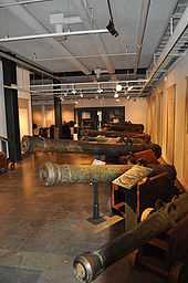 Photograph of an exhibition hall with several types of bronze cannons on naval gun carriages