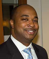 An African-American man wearing a dark suit, white shirt and blue, striped tie