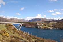 A concrete road bridge spans a body of deep blue water with brown hills beyond