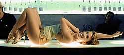 A woman with dark blonde hair is laying on a glass bar counter, which features a blue colour scheme. The woman is dressed in a pale green-tinged jersey top, gold lamé hotpants, and golden high-heels