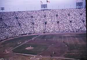 A baseball game being played in a stadium with high stands, seen from the right field side. The United States and Los Angeles flags are flying above the left field foul line, which is shorter than usual
