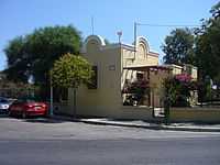 Durrell's house in Rhodes features yellow-painted stucco or plaster walls, with Mediterranean architecture. It is situated alongside an asphalt street, with two cars parked parallel to it. The house is surrounded by several trees, shrubbery, roses and flowering bushes.