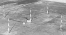 Aerial view of five tall Adcock antenna towers standing on flat terrain; four are arranged in a square, and the fifth one is at the center