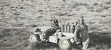 Man standing at the rear of a jeep which is heavily loaded with fuel cans and armed with twin guns at the front