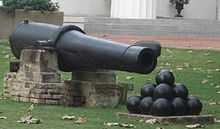 A long 19th-century cannon with a stack of cannon balls in front of it, sitting on a lush green lawn.