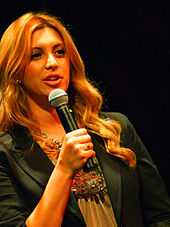 A photograph of a blonde woman speaking into a black microphone which she is holding in her right hand while wearing a black jacket