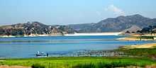 A light blue, broad lake is formed by a dam seen in the distance in a wide valley between arid hills