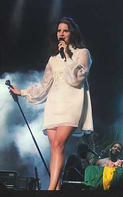 A light-skinned, brown-haired young woman wearing a white long-sleeved dress is standing on a stage singing into a microphone.