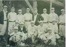 Group of 14 men. The majority are standing, while some in the front are sitting.