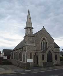 A pale stone building with a tiled roof sloping further on the left than on the right.  A tower with a two-light lancet window, diamond-shaped decoration and a short spire stands in the near corner.  A porch, with two doors and two windows in a lancet style, stands in front of the main body of the church, dominated by a four-light lancet.
