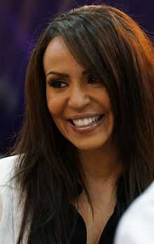 A close-up headshot of a tanned, Caucasian woman with long brown hair, who is wearing a black blouse and white jacket.