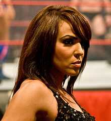 A close-up headshot of a tanned, Caucasian woman with brown hair, slightly past shoulder length, who is wearing a black top.