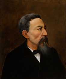 Color portrait of a bearded man wearing a black suit, facing right.