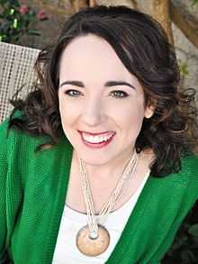 A photograph of a smiling woman wearing red lipstick, a white necklace, a white shirt, and a green sweater looking at the viewer