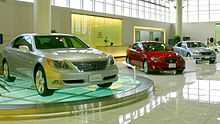 Car showroom displaying three sedans, the nearest on a glass turntable, in front of a reception counter and windows.