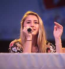 A teenage blonde girl with black floral patterned top, seated behind a table draped in a white cloth looking forward at an unseen audience; a microphone in her right hand is held to her mouth as she gestures with her left hand.