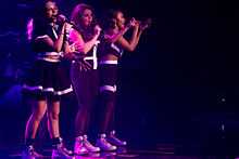 Four women on a stage with microphones in their hands. All four women are in a standing position and are wearing black and white outfits and Dr. Martens footwear as bright purple and dark blue stage lighting shines upon them.