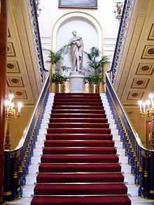 A red-carpeted staircase seen centrally from below with banisters on each side.  At the top is a statue of a standing man wearing a toga and holding a scroll, and on each side are chandeliers on stands